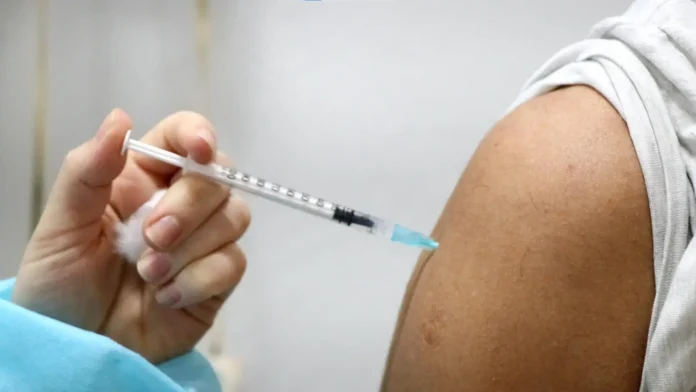 Seringa sendo preparada para aplicação em um braço exposto, com algodão visível ao fundo.