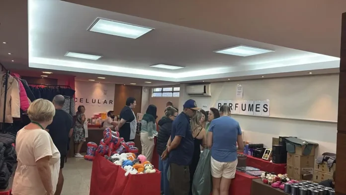 Ambiente interno de um bazar beneficente com pessoas, mesas e produtos como celulares e perfumes expostos.