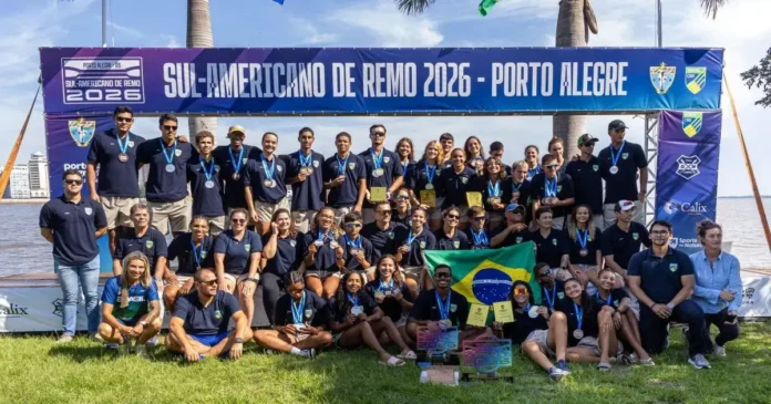 Grupo de pessoas em um evento de remo, exibindo medalhas e troféus, com uma bandeira do Brasil e um banner ao fundo.