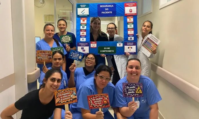 Equipe de saúde segurando placas sobre segurança do paciente em corredor hospitalar. Destaques incluem compromisso com protocolos de segurança e comunicação efetiva.