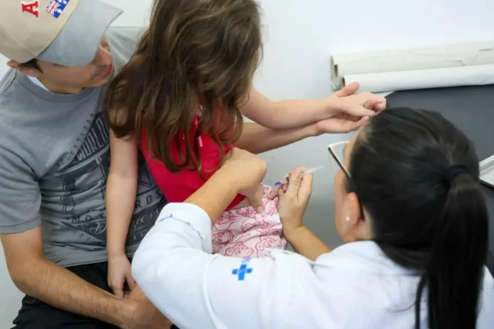 Criança recebendo uma vacina no braço, enquanto um adulto a segura e uma profissional de saúde aplica a injeção.
