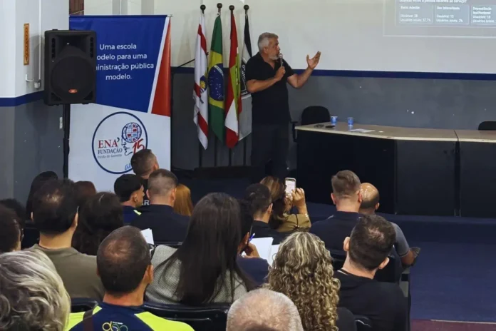 Palestrante em evento da Escola de Governo, com público atento e bandeiras ao fundo, apresentando informações em um telão.