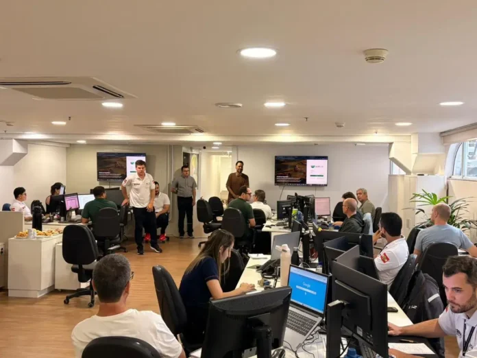 Ambiente de trabalho com várias pessoas em suas mesas, assistindo a uma apresentação em tela grande. Snacks estão disponíveis em uma mesa.