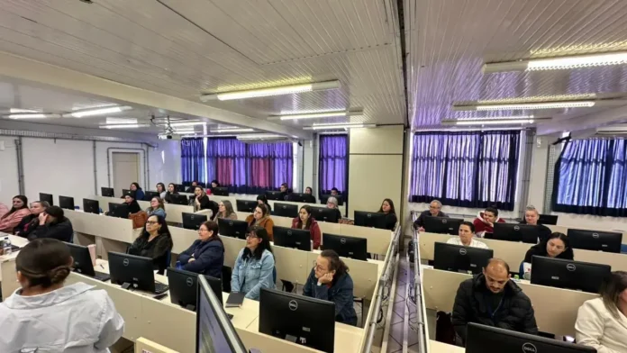 Grupo de pessoas assistindo a uma apresentação em um ambiente com computadores. Cortinas nas janelas e mesas organizadas.