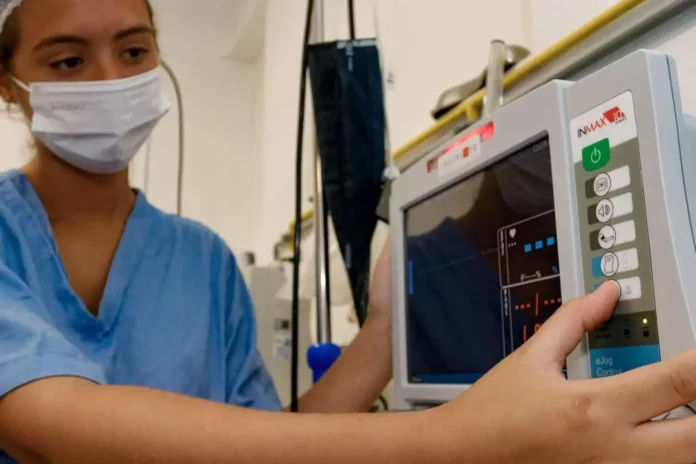 Pessoa com máscara e vestimenta hospitalar operando um monitor médico, ajustando configurações.