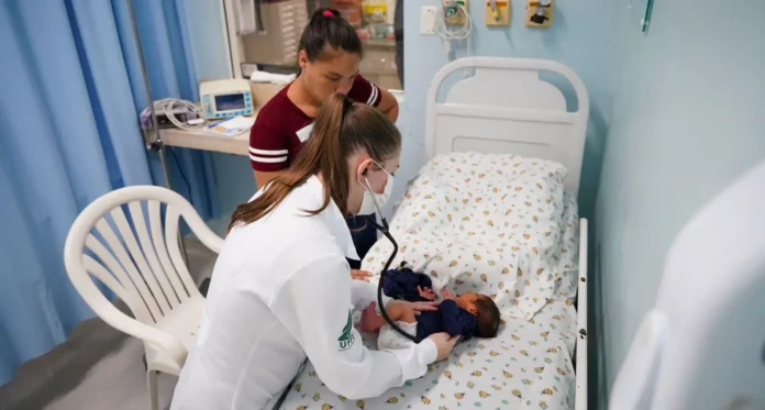 Profissional de saúde examina um recém-nascido em uma cama de hospital, enquanto uma acompanhante observa.