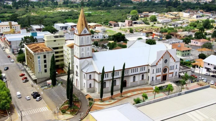 Vista aérea de um edifício localizado em uma área urbana, com vegetação ao fundo e ruas nas proximidades.