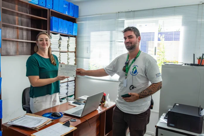 Pessoa entrega um certificado a outra em um escritório com documentos organizados e equipamentos de trabalho visíveis.