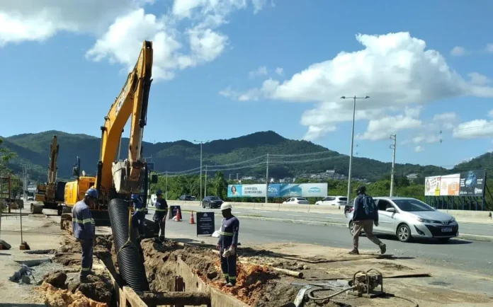 Obras de escavação em via urbana com máquinas e trabalhadores, próximo a montanhas e veículos passando.