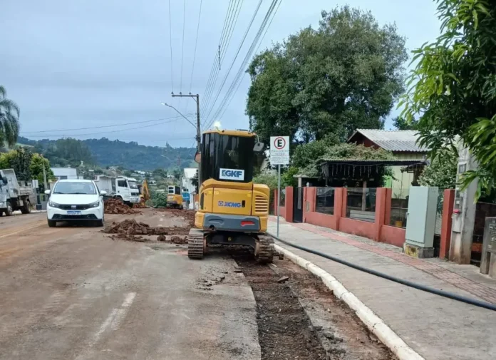 Maquinário de construção em obra na rua, com veículos estacionados e sinalização visível ao fundo.