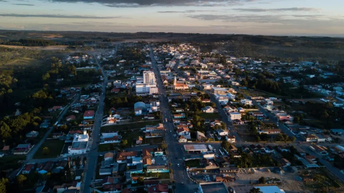 Vista aérea de um município com ruas, casas e prédios, cercado por áreas verdes e colinas ao fundo.
