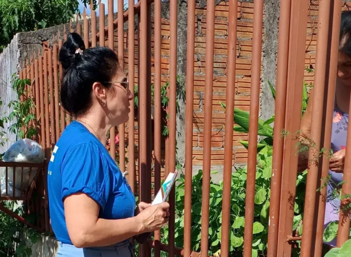 Duas mulheres conversando através de uma grade, em um ambiente ensolarado com plantas ao redor.