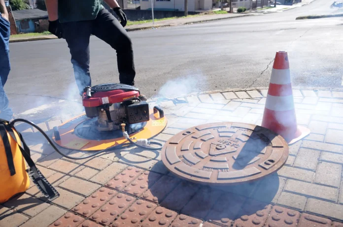 Equipamento de teste de fumaça próximo a um bueiro, com fumaça visível, e cone de sinalização ao lado.