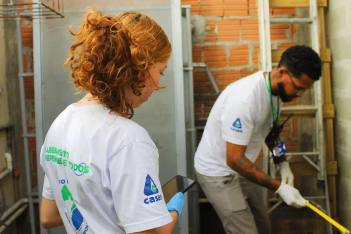 Duas pessoas em camisetas brancas trabalham em um local com estrutura de obras, utilizando equipamentos de medição.