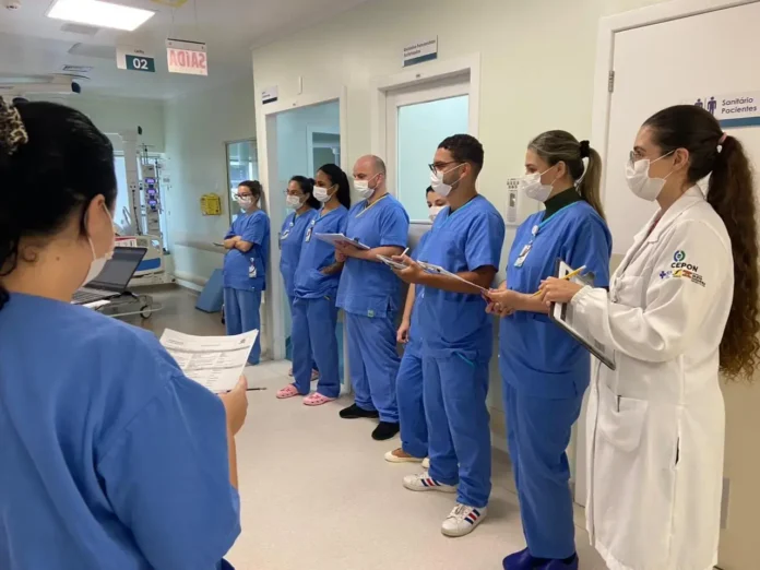 Grupo de profissionais de saúde em uniformes azuis, usando máscaras, escutando alguém à frente e segurando anotações em um hospital.