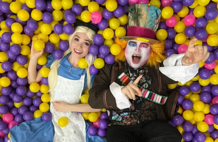 Duas pessoas sentadas em uma piscina de bolinhas coloridas, uma vestida de Alice e outra como Chapeleiro Maluco.