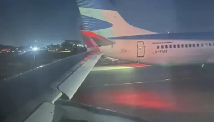 A imagem mostra a asa de um avião à noite, com outro avião próximo, iluminado por luzes na pista.