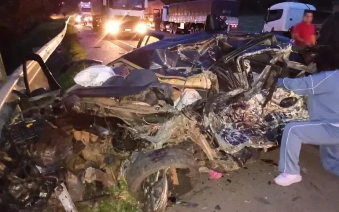Veículo severamente danificado após acidente, com parte da carenagem destruída e socorristas no local. Caminhões em movimento na estrada ao fundo.