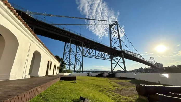 Vista da Ponte Hercílio Luz ao fundo, com canhões e gramado em primeiro plano. céu claro com algumas nuvens.