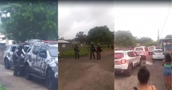 Diversas viaturas da polícia militar estão estacionadas em uma rua com policiais armados e moradores observando.