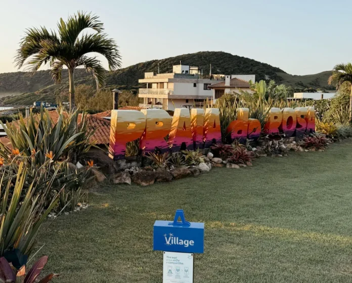 Letreiro colorido com a inscrição "Praia do Rosa" em um ambiente com plantas e paisagem ao fundo.