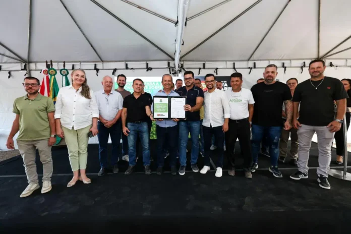 Grupo de pessoas em evento sob uma tenda, apresentando um documento oficial, com bandeiras ao fundo.