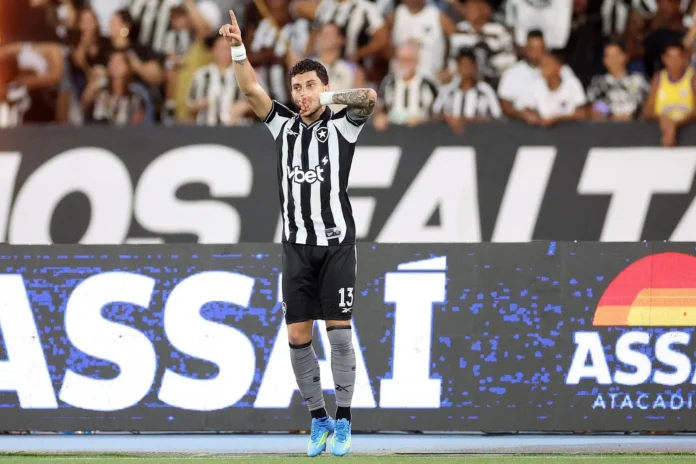 Atleta em campo comemorando, levantando um dedo e fazendo gesto de silêncio, com torcedores ao fundo.