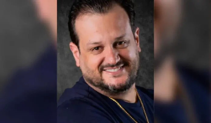 Homem sorridente com cabelo curto, usando camiseta escura e corrente no pescoço. Fundo cinza.