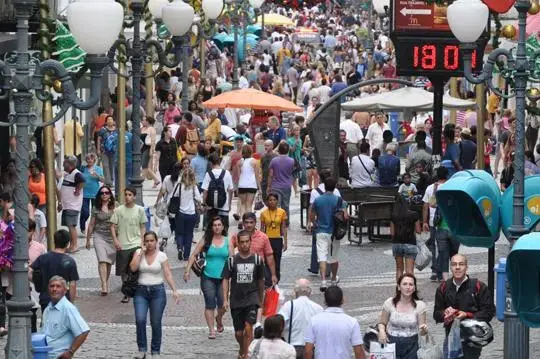 Rua movimentada com muitas pessoas caminhando, bancas de comércio e um relógio digital indicando 18:01.
