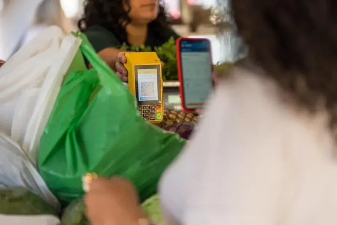 Máquina de pagamento sendo utilizada em uma feira, com sacolas de compras ao fundo e um celular visível.