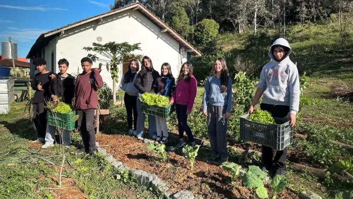 Grupo de jovens em um jardim, segurando caixas com hortaliças, ao fundo uma casa e vegetação.