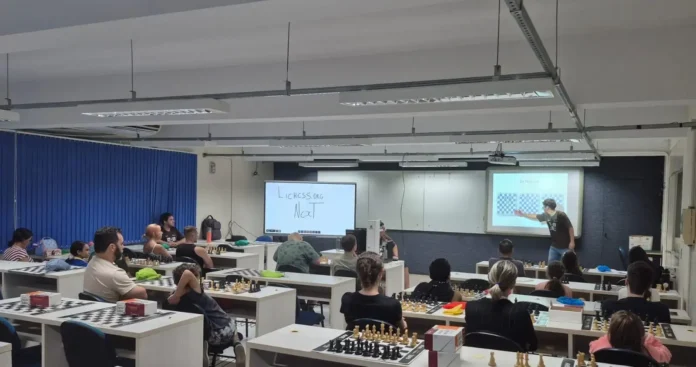 Sala de aula com pessoas assistindo a uma apresentação sobre xadrez; quadro branco com texto "lichess.org" e "Next".