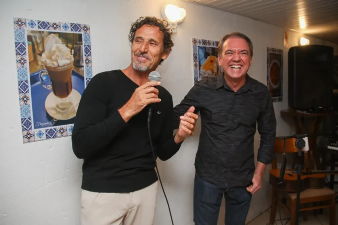 Duas pessoas se divertem em um ambiente interno, uma delas segura um microfone enquanto ri. Algumas fotos decoram a parede ao fundo.