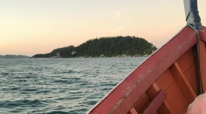 Borda de um barco com vista para uma ilha ao fundo, durante o pôr do sol em um mar calmo.