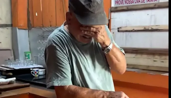 Homem com chapéu cobrindo a face, de camisa cinza, em ambiente interno com objetos ao fundo.
