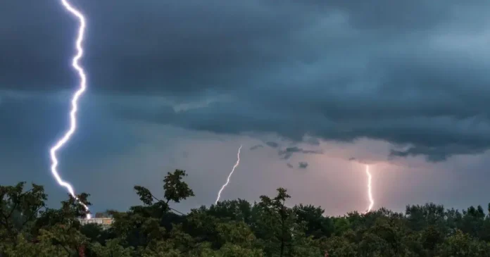 Relâmpagos iluminam um céu nublado, com nuvens escuras e árvores ao primeiro plano.