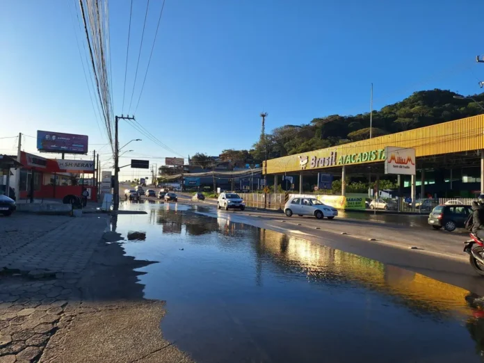 Rua alagada com veículos trafegando, lojas e céu claro ao fundo. Sinalização visível.