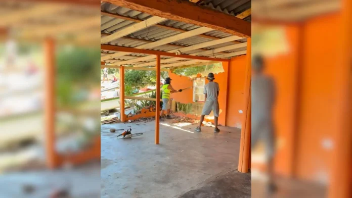 Dois trabalhadores demolem uma parede de um espaço interno, cercados por lixo e ferramentas.