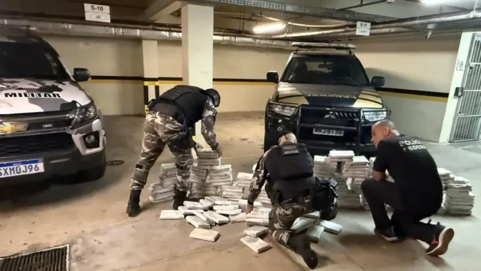 Agentes de segurança organizam pacotes de drogas em um estacionamento, com veículos da polícia ao fundo.