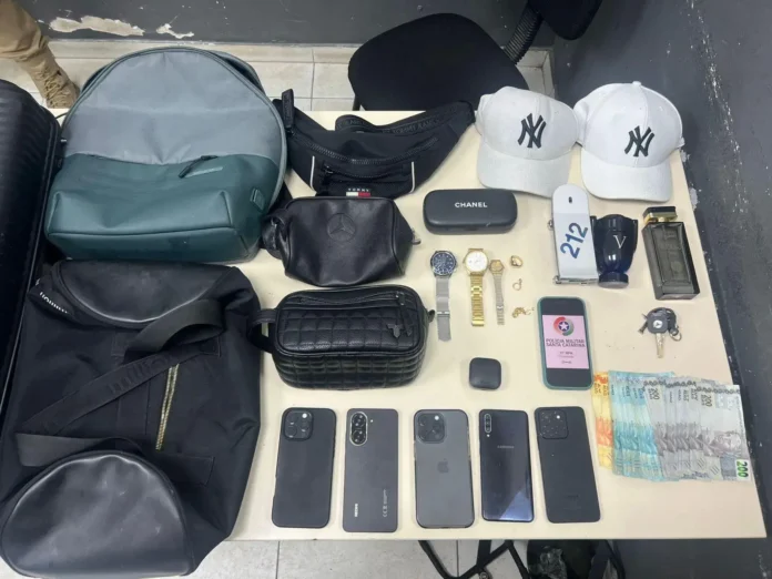 Diversos objetos apreendidos, incluindo bolsas, relógios, celulares, um frasco de perfume, dinheiro em notas e bonés.