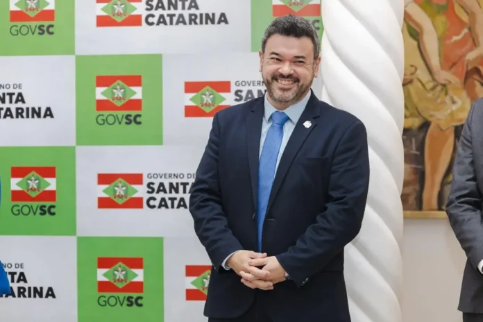 Homem sorridente em um evento com fundo do Governo de Santa Catarina, com logotipo e bandeira ao fundo.