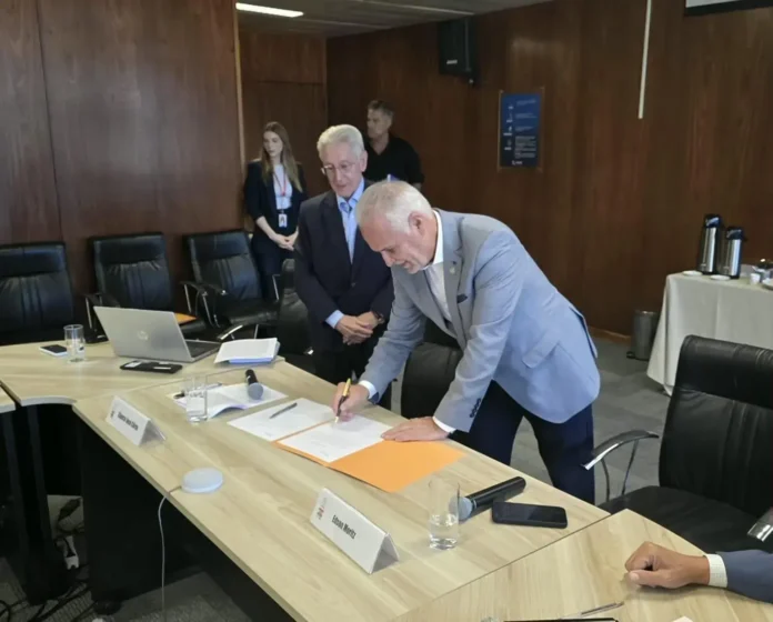 Pessoa assinando documento em mesa de reunião, com laptop e copos de água visíveis ao fundo.