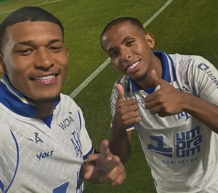 Dois jogadores de futebol sorrindo e fazendo sinal de positivo, usando camisas brancas com detalhes azuis, em campo.