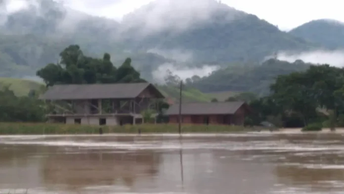 Paisagem com casas parcialmente submersas em área alagada, cercada por montanhas e neblina.