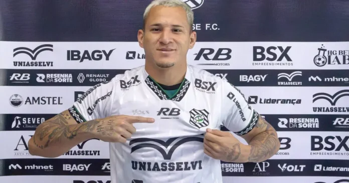 Jogador apresenta a camiseta do time, com logotipos de patrocinadores e fundo neutro.