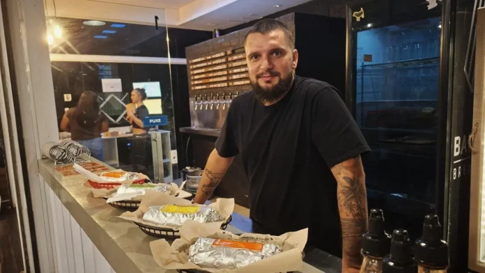 Homem com barba e tatuagens em um balcão, ao fundo, interage com uma mulher. Vários pratos embrulhados estão sobre a bancada.