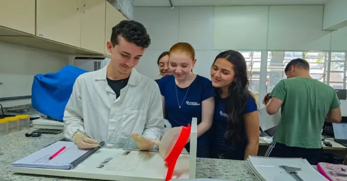 Estudantes em laboratório observando um experimento, com equipamento e materiais de pesquisa visíveis.