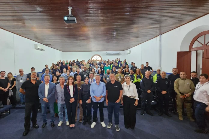 Grupo de pessoas em um auditório, com alguns em uniforme, assistindo a uma apresentação sob luzes e projeção.