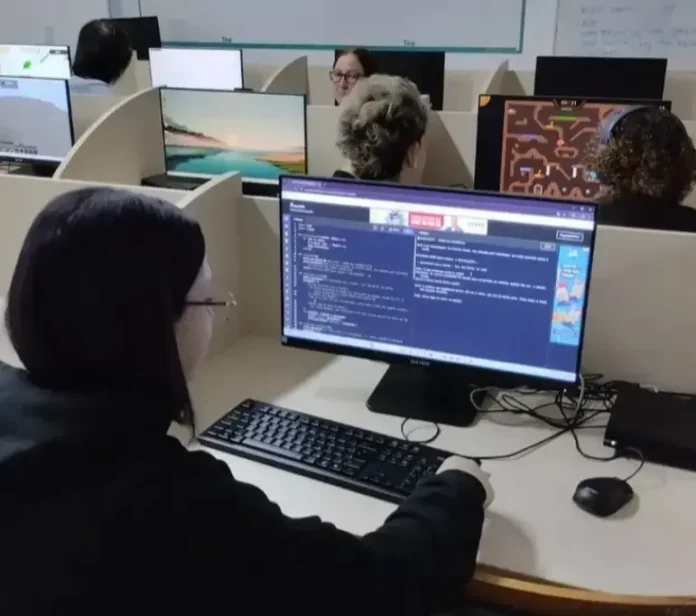 Aluno trabalhando em computador, digitando código em tela com outras pessoas ao fundo.