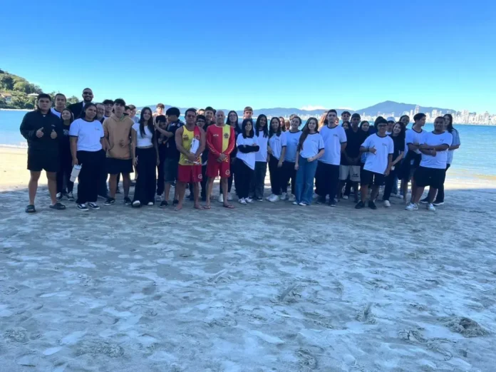 Grupo de jovens em uma praia, posando em frente ao mar e à cidade ao fundo.